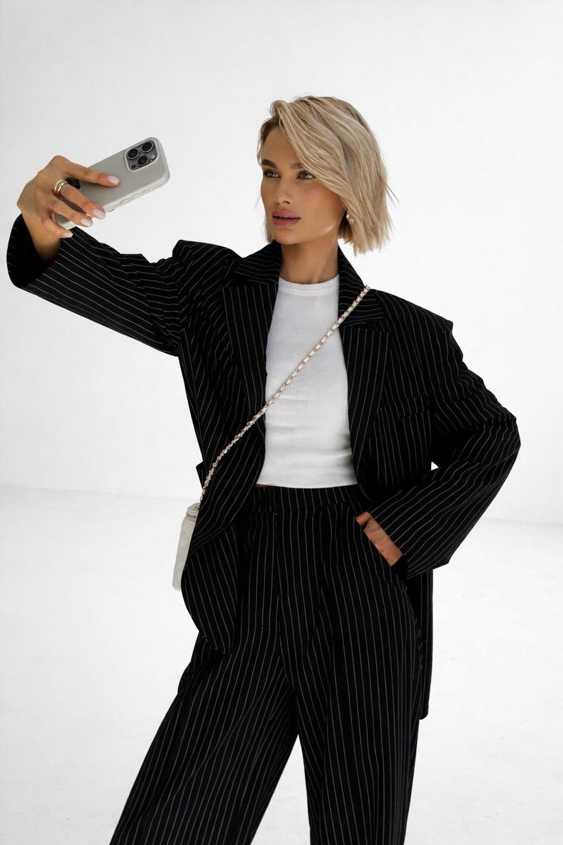 May include: Black pinstripe blazer and matching pants. The blazer has a classic notched lapel and a two-button closure. The pants are high-waisted with a straight leg. A white top is worn underneath the blazer. A small white purse with a chain strap is worn over the shoulder.