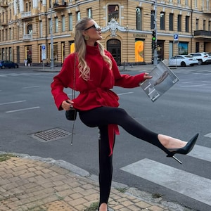 May include: A vibrant red long-sleeved wrap jacket with a high collar and a tie-waist belt, paired with black stirrup leggings and black pointed-toe high-heeled pumps. A small black shoulder bag with a chain strap and a magazine are also visible.