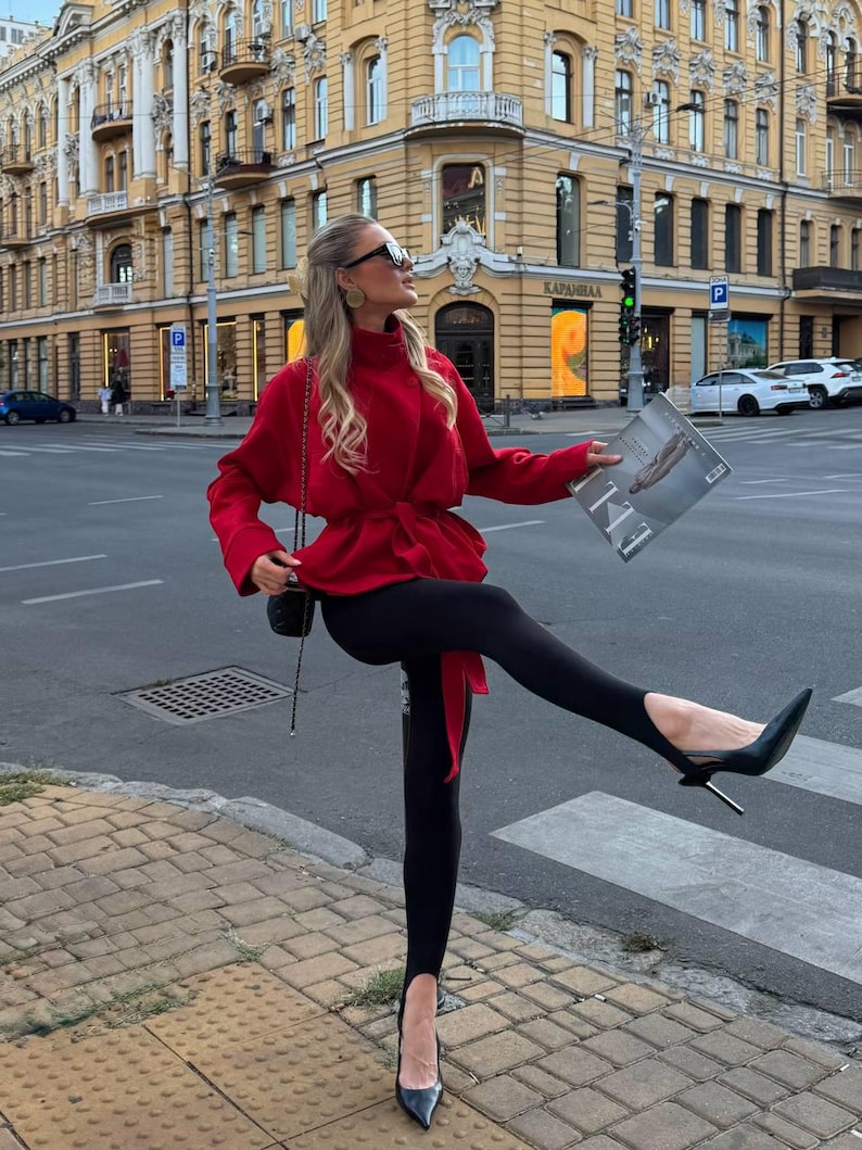May include: A vibrant red long-sleeved wrap jacket with a high collar and a tie-waist belt, paired with black stirrup leggings and black pointed-toe high-heeled pumps. A small black shoulder bag with a chain strap and a magazine are also visible.