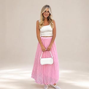 May include: A white sleeveless crop top paired with a long, pleated, light pink skirt. The outfit is accessorized with a small, structured white handbag featuring a top handle and a pair of white, closed-toe, flat shoes with a strap across the top.