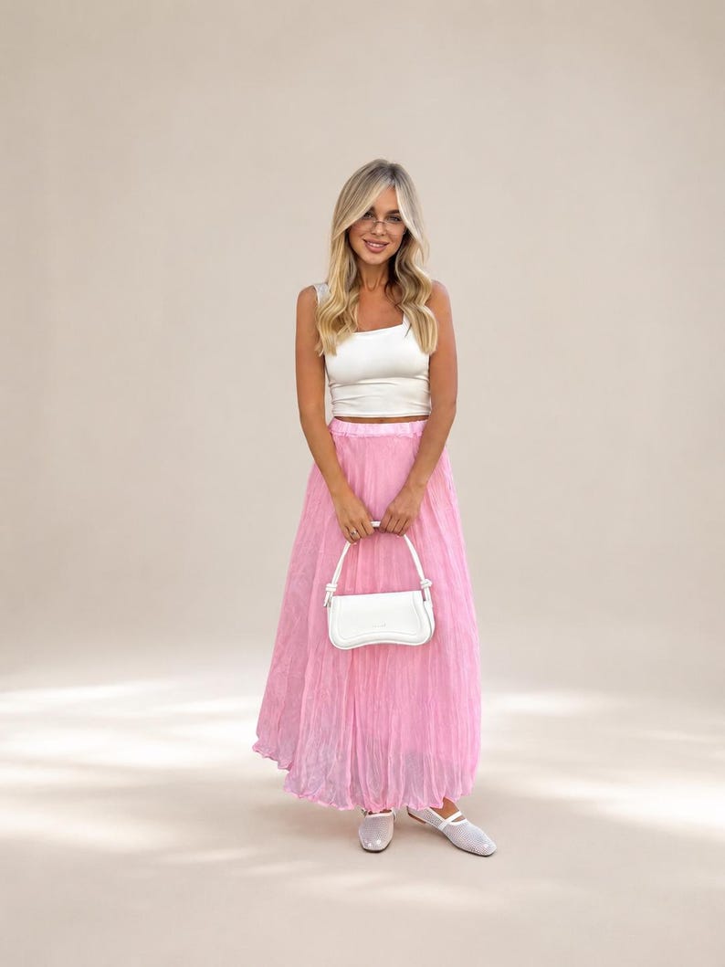May include: A white sleeveless crop top paired with a long, pleated, light pink skirt. The outfit is accessorized with a small, structured white handbag featuring a top handle and a pair of white, closed-toe, flat shoes with a strap across the top.