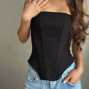 May include: A black strapless corset top with vertical seams and a textured appearance. The top is paired with light blue denim jeans. The overall style is modern and chic.