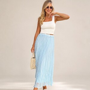 May include: A white sleeveless square-neck crop top paired with a light blue pleated maxi skirt. The outfit includes a small woven straw handbag and white mesh-textured flat shoes. The look is completed with dark-tinted sunglasses.