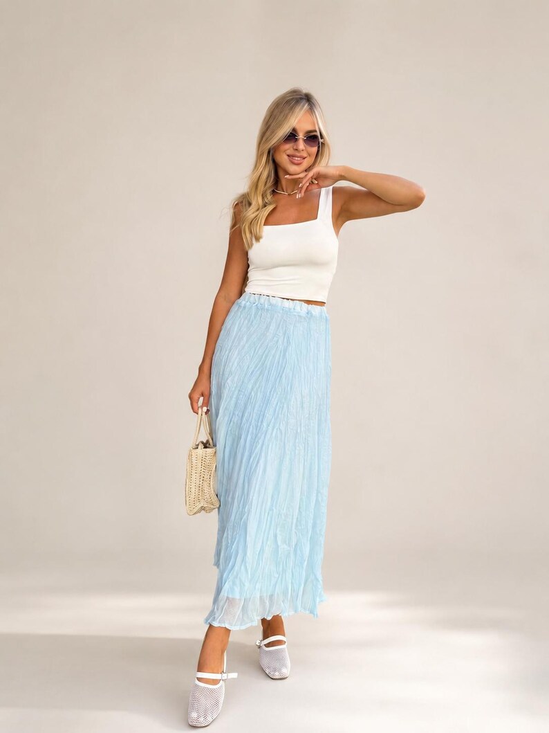 May include: A white sleeveless square-neck crop top paired with a light blue pleated maxi skirt. The outfit includes a small woven straw handbag and white mesh-textured flat shoes. The look is completed with dark-tinted sunglasses.