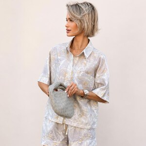 Puede incluir: Conjunto de camisa y pantal&oacute;n corto de lino gris claro con estampado floral. La camisa tiene mangas cortas y cierre de botones. Se sostiene un peque&ntilde;o bolso de mano gris tejido. El conjunto se complementa con un reloj plateado.