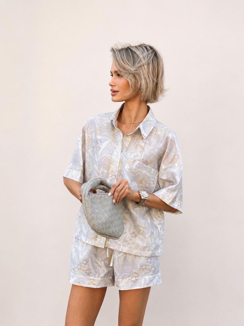 Puede incluir: Conjunto de camisa y pantal&oacute;n corto de lino gris claro con estampado floral. La camisa tiene mangas cortas y cierre de botones. Se sostiene un peque&ntilde;o bolso de mano gris tejido. El conjunto se complementa con un reloj plateado.