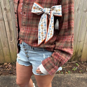 May include: A red and brown plaid button-up shirt with a white and floral patterned bow. The shirt is paired with light blue denim shorts with distressed details. White boots complete the outfit.