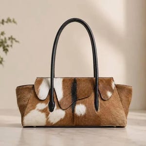 Könnte beinhalten: Eine rechteckige Handtasche mit braun-weißem Kuhfellmuster und einer texturierten Oberfläche aus Fell. Die Tasche verfügt über zwei schmale schwarze Ledergriffe an der Oberseite, einen Klappverschluss und eine strukturierte, längliche Form.