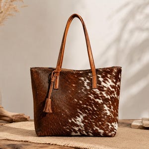 May include: A tote bag featuring a brown and white cowhide pattern with a textured hair-on-hide finish. The bag includes two thin tan leather shoulder straps and a matching leather tassel charm attached to one side of the top handle base.