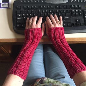 May include: A pair of vibrant red crocheted arm warmers. The ribbed texture of the warmers is visible, and they extend from the wrists to the mid-forearm. The warmers are worn while typing on a black keyboard.