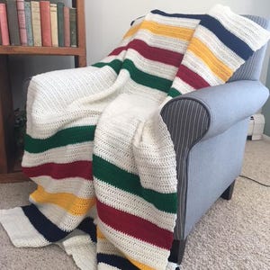 以下が含まれることがあります： クリーム色の、ネイビー、イエロー、レッド、グリーンの大胆なストライプが入ったかぎ針編みのブランケットが、グレーのアームチェアにかけられています。ブランケットの質感がはっきりと見え、手作りの品質を示しています。配色はクラシックなデザインを彷彿とさせます。