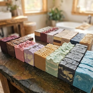 May include: A selection of handmade soaps in a variety of colours and designs. The soaps are displayed on a dark, textured surface. Colours include shades of purple, pink, green, blue, and brown. Some soaps have decorative toppings.