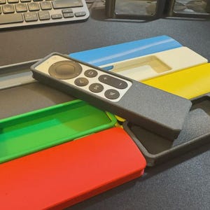 May include: A dark gray remote control with a circular navigation pad and several buttons, resting on a collection of rectangular protective cases in various colors including red, green, yellow, white, blue, and dark gray, each with a cutout.