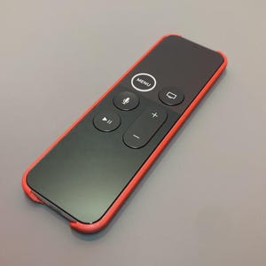 May include: A black remote control encased in a vibrant red silicone cover. The remote features a circular navigation pad with a central MENU button, a microphone icon, a television icon, a play-pause button, and volume plus and minus controls.