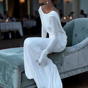 May include: A white, one-shoulder gown with a long sleeve and a flowing skirt. The dress is worn by a person seated on a teal velvet chaise longue. The background is a blurred interior with soft lighting.