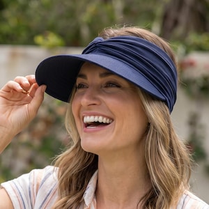 Può includere: Una visiera parasole blu navy caratterizzata da un'ampia tesa curva e da un dettaglio in tessuto arricciato e intrecciato sulla parte anteriore. La visiera è realizzata in un materiale flessibile a tinta unita con un design plissettato.