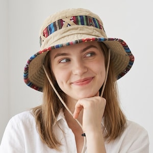 Może przedstawiać: Beżowy kapelusz typu bucket hat z szerokim rondem, ozdobiony kolorową, wzorzystą taśmą wokół główki oraz na spodniej stronie ronda. Kapelusz posiada jasny, regulowany pasek pod brodę z przesuwanym stoperem dla zapewnienia stabilnego dopasowania.