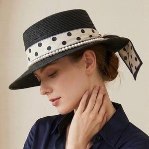 Puede incluir: Un sombrero de paja negro con copa plana y ala ancha. El sombrero está adornado con una cinta de color crema con lunares negros y una hilera de perlas artificiales blancas que rodean la base de la copa, atada en un lazo en el lateral.