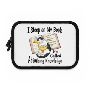 Op de afbeelding: Een witte laptoptas met een zwarte rand en rits. De tas toont een cartoonkat die op een boek slaapt en een gele eend knuffelt. De tekst op de tas luidt "I Sleep on My Book" en "It's Called Absorbing Knowledge."
