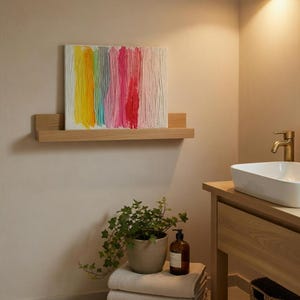 May include: An abstract artwork with vertical brushstrokes in yellow, green, grey, pink, and red. The painting is displayed on a wooden shelf. The bathroom scene includes a sink, towels, and a potted plant.