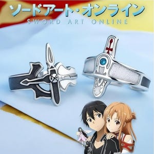 Puede incluir: Dos anillos plateados con diseños intrincados. Uno presenta un emblema de espada en blanco y negro, el otro una cruz roja y una gema azul. Los anillos se muestran con personajes de anime y el texto "SWORD ART ONLINE".
