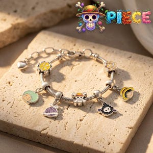 Op de afbeelding: Een zilverkleurige bedelarmband met diverse kleurrijke emaille bedels geïnspireerd op One Piece, waaronder een strooien hoed, een schedel-embleem, een paarse vrucht en karaktergezichten, met een hartvormige sluiting en een ketting.