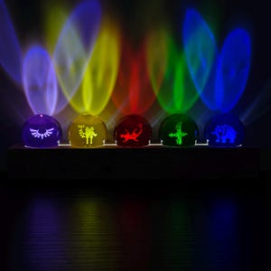 Puede incluir: Cinco esferas de cristal dispuestas en fila sobre una base rectangular de madera. Cada esfera brilla en un color diferente—morado, amarillo, rojo, verde y azul—y presenta un símbolo blanco grabado único, proyectando haces de luz.