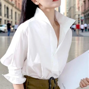 May include: A white button-down shirt with a wide, structured collar and long sleeves. The shirt is worn with olive green trousers and a black belt. The background shows a city street with blurred figures.