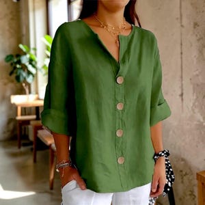 Puede incluir: Una camisa verde de lino con cuello en V y mangas tres cuartos. La camisa tiene botones grandes y redondos de madera en la parte delantera. Las mangas están enrolladas. La camisa se combina con pantalones blancos.