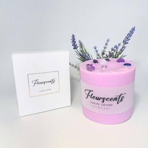 Puede incluir: Una vela de lavanda rosa claro con el texto "Fleurscents" y "Luxury candle" en una etiqueta blanca. La vela está decorada con cristales y una ramita de lavanda. A la izquierda hay una caja blanca con un patrón geométrico y el logotipo de Fleurscents.