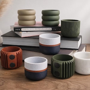 Può includere: Una collezione di fioriere decorative in ceramica di vari colori e design. Le fioriere includono un design impilato verde oliva, un design verde scuro strutturato e un design bicolore bianco e blu navy. Le fioriere sono disposte su una superficie di legno.