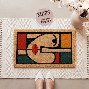 May include: A rectangular coir doormat featuring a stylized abstract face design with bold black outlines. The graphic includes geometric blocks of red, orange, and dark green. The face has a single prominent eye and red lips. The mat has a natural tan texture.