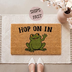 May include: A rectangular doormat made of natural coir fiber, featuring the text "HOP ON IN" in black letters. A cartoon frog in green with orange eyes is waving. The mat is placed on a white patterned rug.