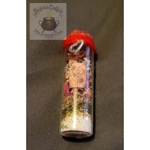 May include: A clear glass vial filled with dried herbs, flowers, and white granules. The vial is topped with a red knitted cap and a silver heart charm. A cork with a tree design is visible. The vial is labeled with the text "Brolivia's Cauldron".