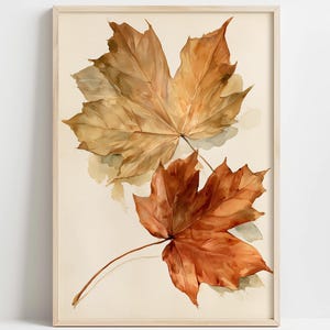 May include: A framed wall art print featuring a watercolour illustration of two maple leaves. The top leaf is a light tan and golden brown, while the bottom leaf is a deeper, vibrant orange-red. Both leaves are set against a cream background.
