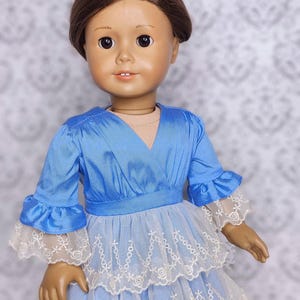 May include: A doll with brown hair wearing a light blue dress with white lace trim. The dress has a layered skirt and ruffled sleeves. The doll's face is detailed with painted features.