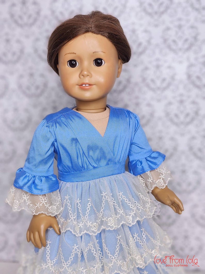 May include: A doll with brown hair wearing a light blue dress with white lace trim. The dress has a layered skirt and ruffled sleeves. The doll's face is detailed with painted features.