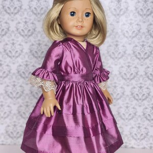 May include: A doll wearing a purple dress with a layered skirt and ruffled sleeves. The dress has a fitted waist and is made of a shiny fabric. The doll has blonde hair, blue eyes, and freckles. The doll is wearing black shoes.