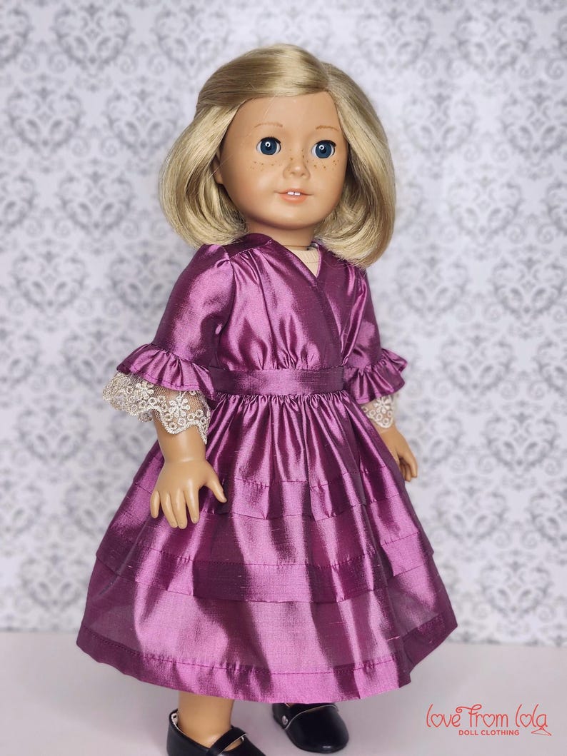 May include: A doll wearing a purple dress with a layered skirt and ruffled sleeves. The dress has a fitted waist and is made of a shiny fabric. The doll has blonde hair, blue eyes, and freckles. The doll is wearing black shoes.