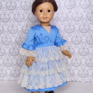 May include: A doll wearing a blue dress with white lace trim and black shoes. The dress has a V-neck and ruffled sleeves. The doll has brown hair and is standing in front of a patterned background. The text "love from lola doll clothing" is in the bottom right corner.