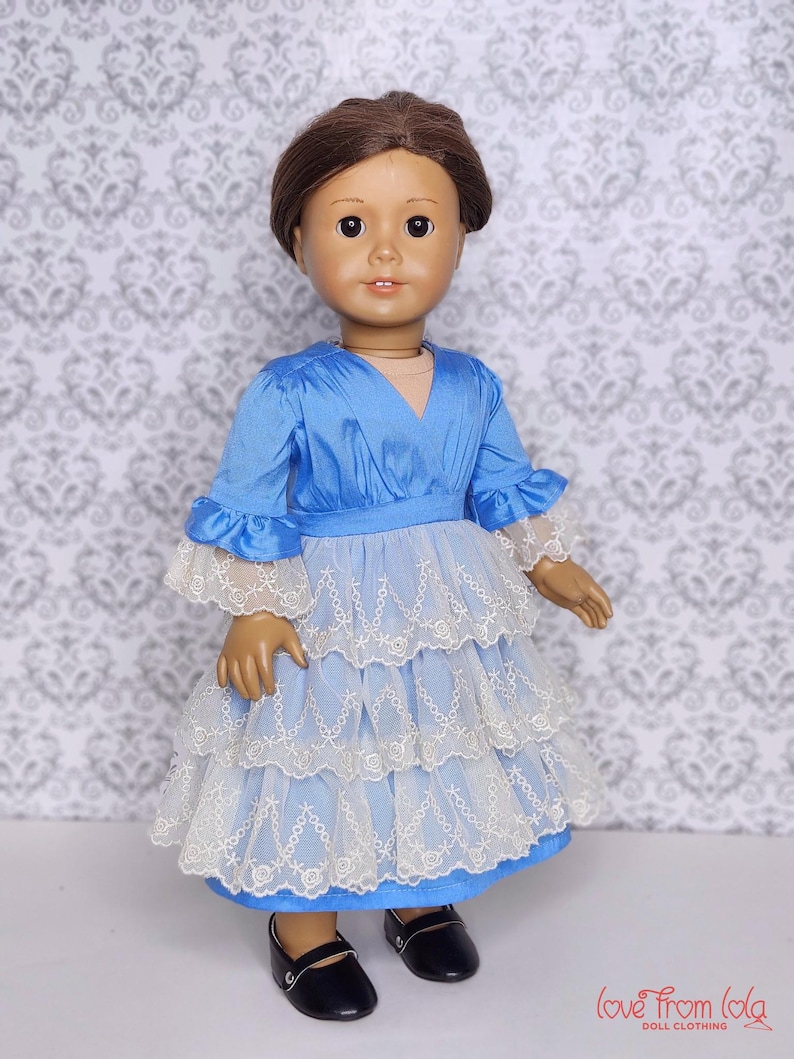 May include: A doll wearing a blue dress with white lace trim and black shoes. The dress has a V-neck and ruffled sleeves. The doll has brown hair and is standing in front of a patterned background. The text "love from lola doll clothing" is in the bottom right corner.