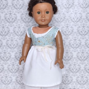 May include: A doll wearing a light blue and white floral print top with a white skirt and shorts. The doll has brown hair and green eyes. The outfit is tied at the waist with a white ribbon. The doll clothing is from Love From Lola.
