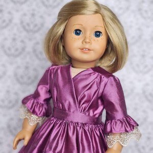 May include: A doll with blonde hair and blue eyes wearing a plum-colored dress. The dress features a wrap-style bodice, ruffled sleeves with lace trim, and a tiered skirt. The doll's clothing is from Love From Lola Doll Clothing.