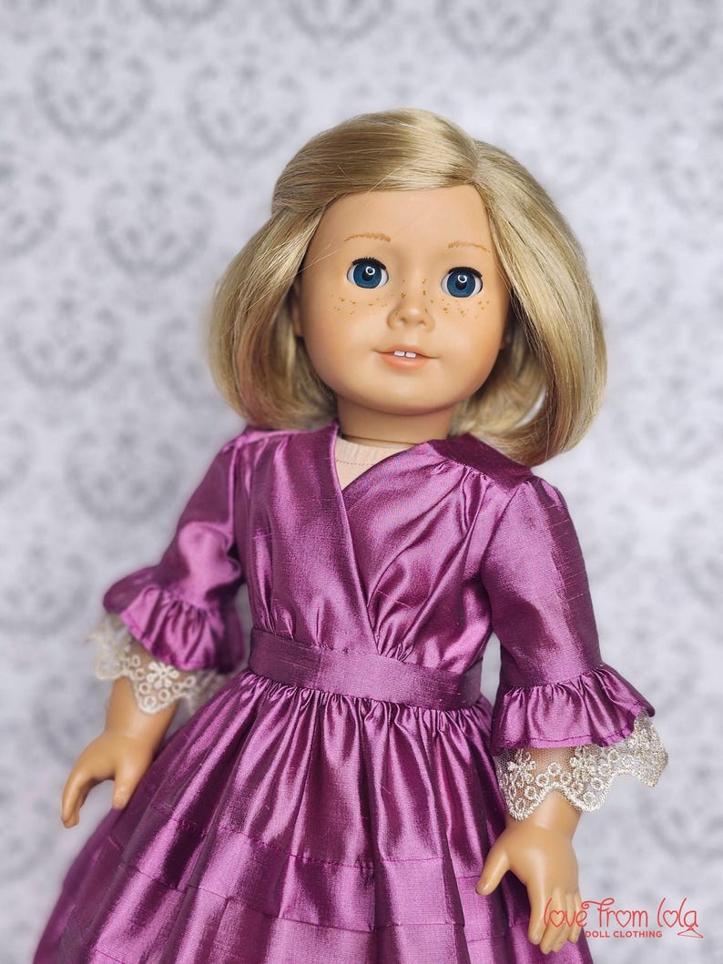 May include: A doll with blonde hair and blue eyes wearing a plum-colored dress. The dress features a wrap-style bodice, ruffled sleeves with lace trim, and a tiered skirt. The doll's clothing is from Love From Lola Doll Clothing.