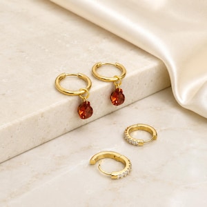 May include: A set of four gold-toned hoop earrings. Two hoops feature a dangling, teardrop-shaped orange gemstone charm. The other two hoops are smaller and encrusted with a row of clear, sparkling stones along the outer edge of the metal band.