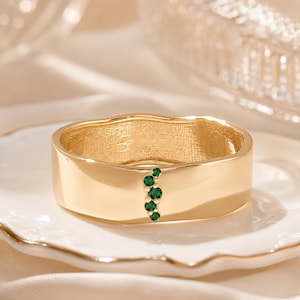 May include: A gold band ring featuring a smooth, polished surface with an irregular, organic edge. A vertical cluster of five small, round, vibrant green gemstones is set into the center of the band, creating a distinct focal point on the metal.
