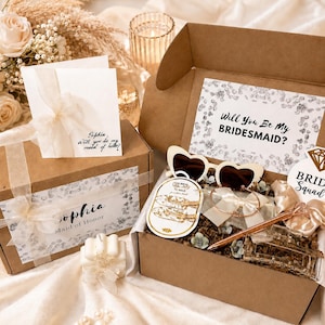 May include: Two brown cardboard boxes with white floral accents and ribbons. One box is open, revealing heart-shaped sunglasses, a pen, and other gifts. The other box has a card that says, "Sophia, will you be my maid of honor?"