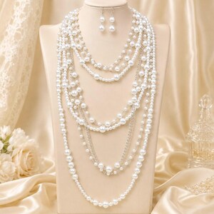 May include: A multi-strand necklace featuring white faux pearls and silver-toned metal chains, arranged in cascading layers. The set includes a pair of matching drop earrings, each consisting of a single white faux pearl suspended from a silver-toned hook.