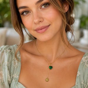 May include: A gold-toned lariat necklace featuring a small, vibrant green heart-shaped pendant and a circular gold disc charm engraved with the name Luna, suspended from a delicate chain that rests against the collarbone area.