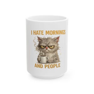 May include: White ceramic coffee mug with the text "I HATE MORNINGS AND PEOPLE". The mug features a grumpy cartoon cat holding a coffee cup, with steam rising from the cup. The cat has gray fur and a stern expression.
